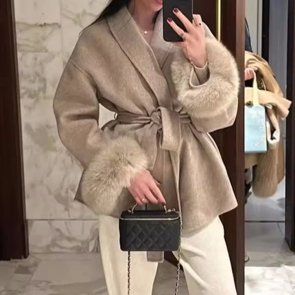 Person wearing a beige coat with fur trim, holding a black handbag, in a marble interior setting.