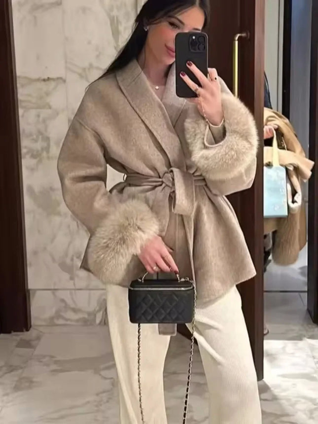 Person wearing a beige coat with fur trim, holding a black handbag, in a marble interior setting.