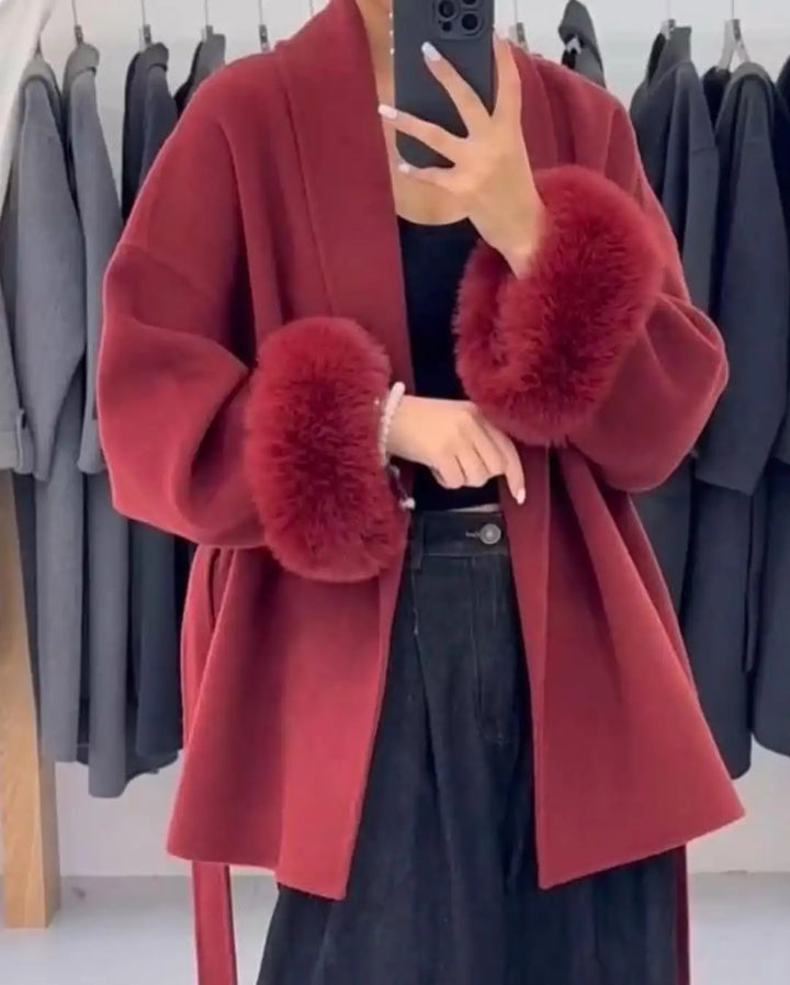 Red coat with fur cuffs worn by a person, with a background of hanging clothes.