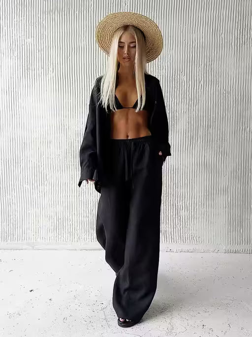 Person wearing a black outfit and straw hat against a white textured wall.