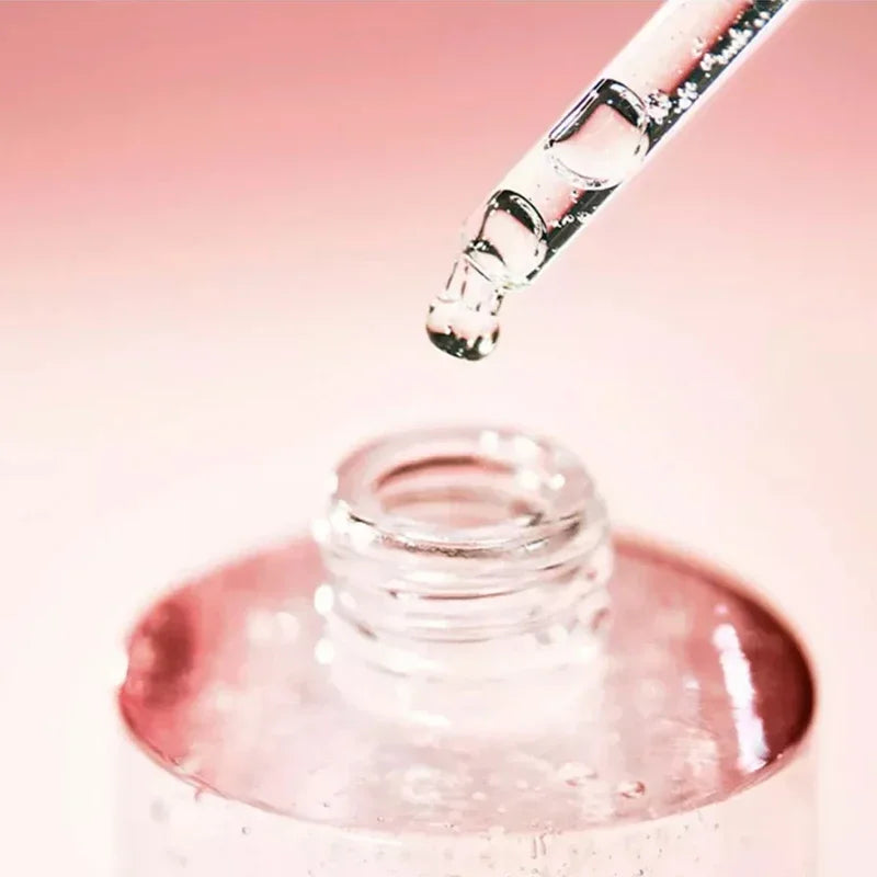 Glass dropper with clear liquid over a small glass container on a pink background