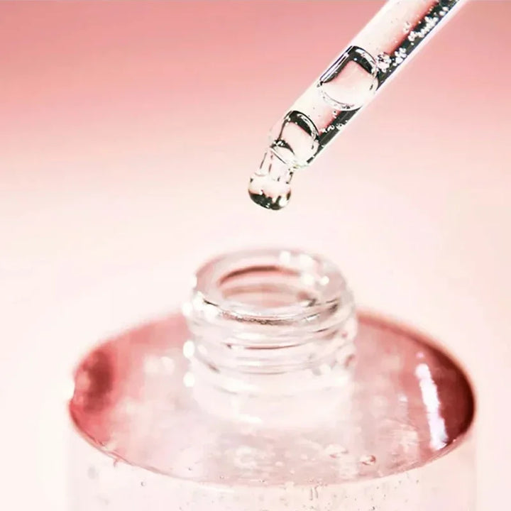 Glass dropper with clear liquid over a small glass container on a pink background