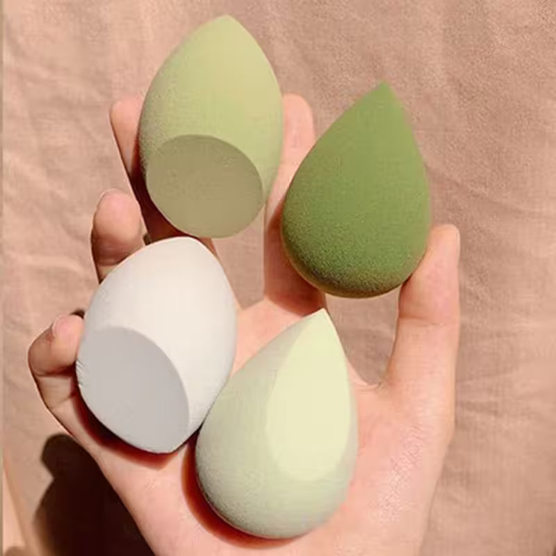 Hand holding four green and gray makeup sponges on a beige background