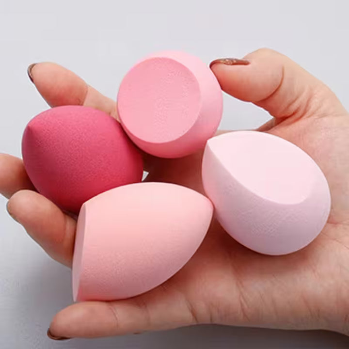 Hand holding four pink makeup sponges against a light gray background